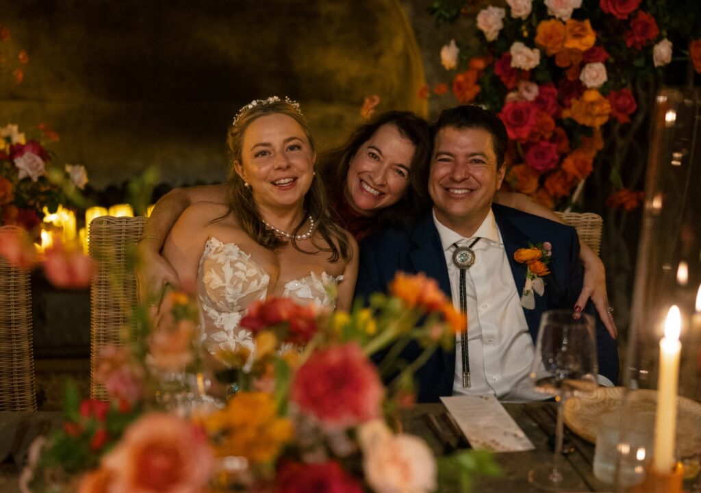 A bride in a floral lace gown and a groom in a blue suit with a bolo tie smile with a guest at their candlelit wedding reception at Villa Santa Cruz.