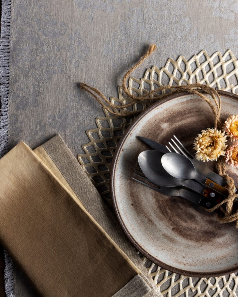 A rustic table setting featuring a Chilewich Dahlia lace placemat, a hand-thrown ceramic plate, and Kim Seybert dip-dyed napkins tied with natural twine and dried flowers.