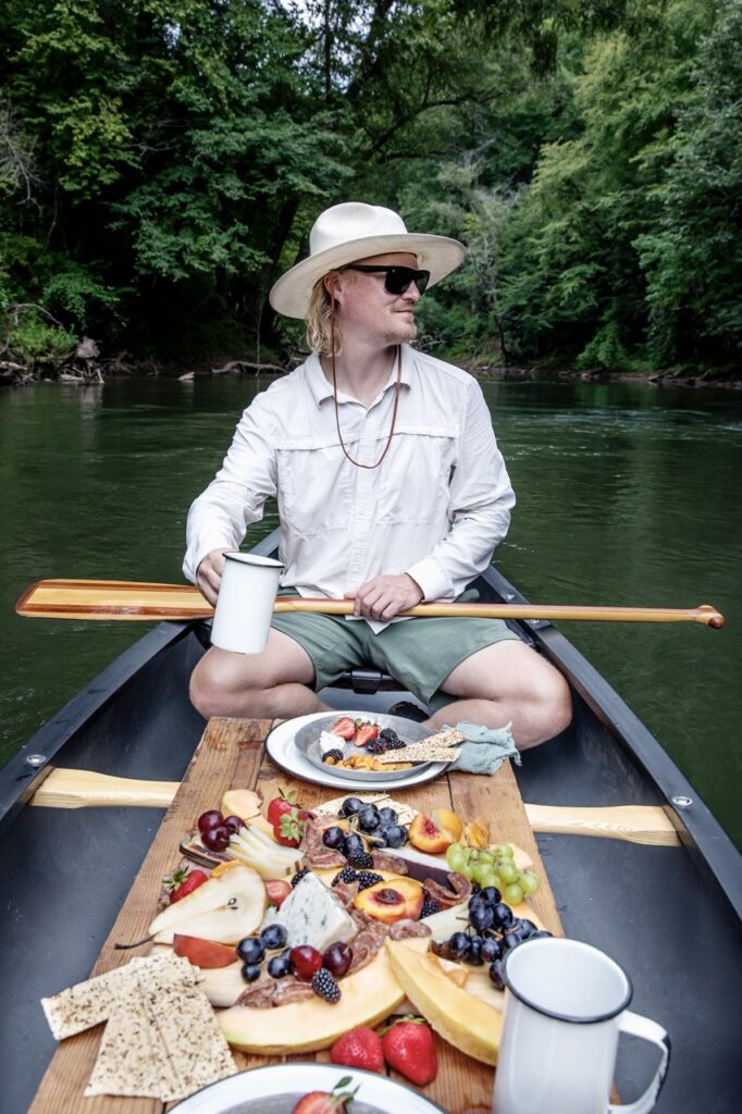 A man sits in a canoe with a charcuterie spread in front of him.