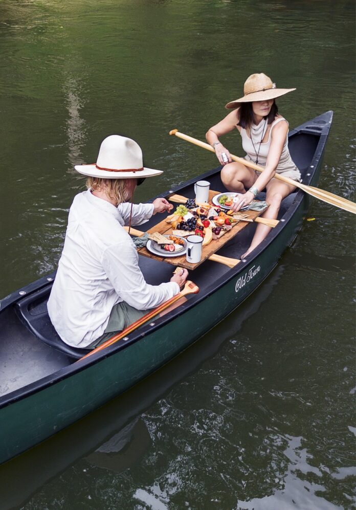 A man and woman in hats sit in a canoe with a charcuterie plate between them.