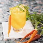 A Carrot Moscow Mule in a yellow color sits on a white cloth with a carrot leaning against it.