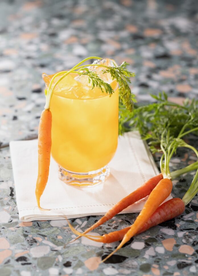 A Carrot Moscow Mule in a yellow color sits on a white cloth with a carrot leaning against it.