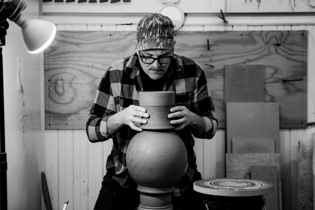 Ceramist Jeff Schwarz in his Pittsburgh studio, holding a large contemporary ceramic vessel with textured surfaces and luxury fashion logo motifs.