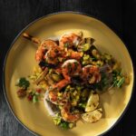 A yellow plate of char grilled shrimp skewers with blue corn grits and golden beet salad under it and slices of charred limes on the sides.