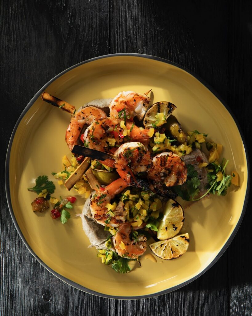 A yellow plate of char grilled shrimp skewers with blue corn grits and golden beet salad under it and slices of charred limes on the sides.