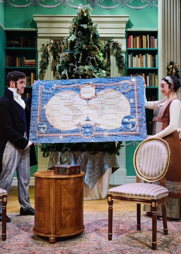 A man and woman in costume hold up a quilt on stage in front of a Christmas Tree.