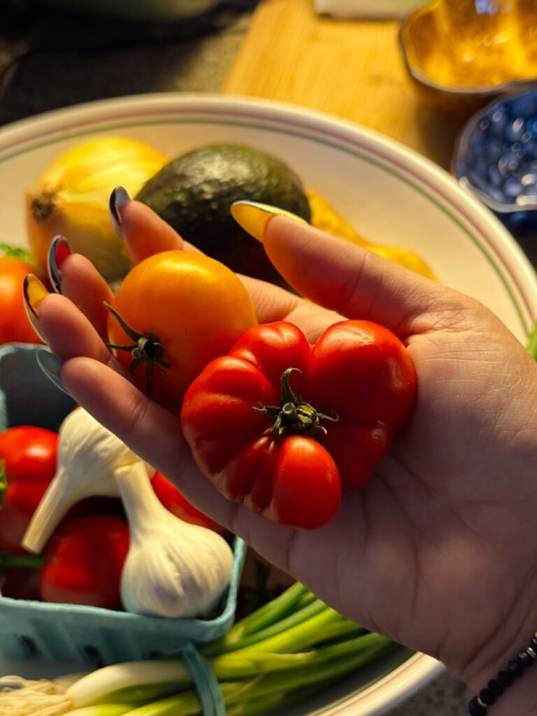 A person holds a red and orange tomato over a bowl of veggies.