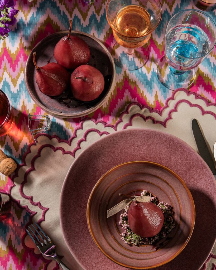 A purple plate with a purple pear inside all sitting on a pattern tablecloth.
