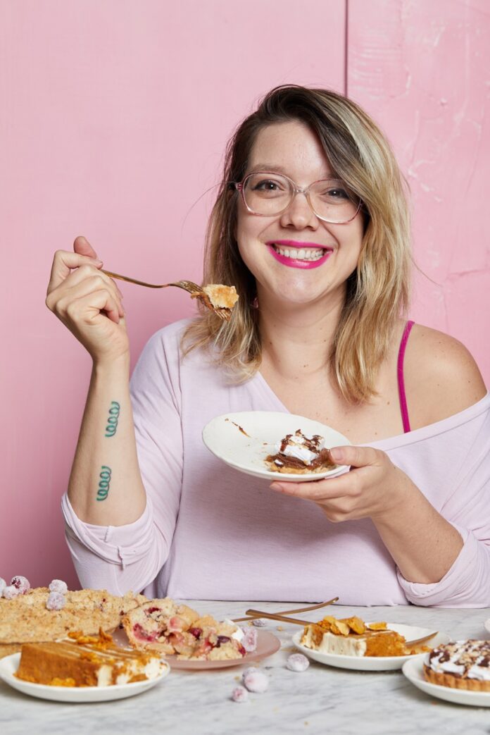 Casey Renee eats cake off a white plate against a pink backgrounf.