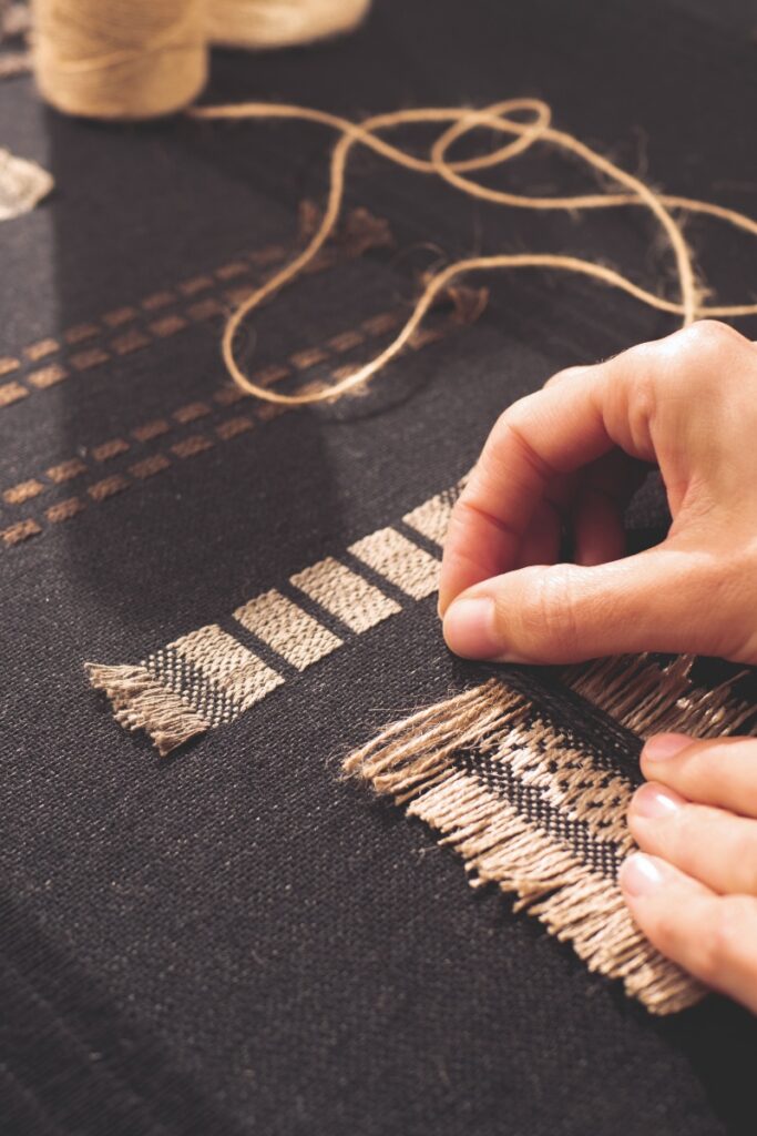 An artisan's hands meticulously weaving a textured Elitis wall covering that combines raffia, linen, and wool in an intricate, abstract pattern inspired by the Japanese concept of yūgen.