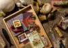 Artisan Crafted Pieces for Home and Fashion A creative mood board by Danny Mankin featuring a collection of vintage books, personal sketches, gold jewelry, and varied fabric textures laid out on a burlap surface.