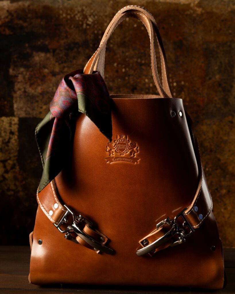 Handcrafted Michael Noelle Soigné Tote in cream leather with an orange interior, styled on a wooden stool next to an orange armchair and a glass of champagne.