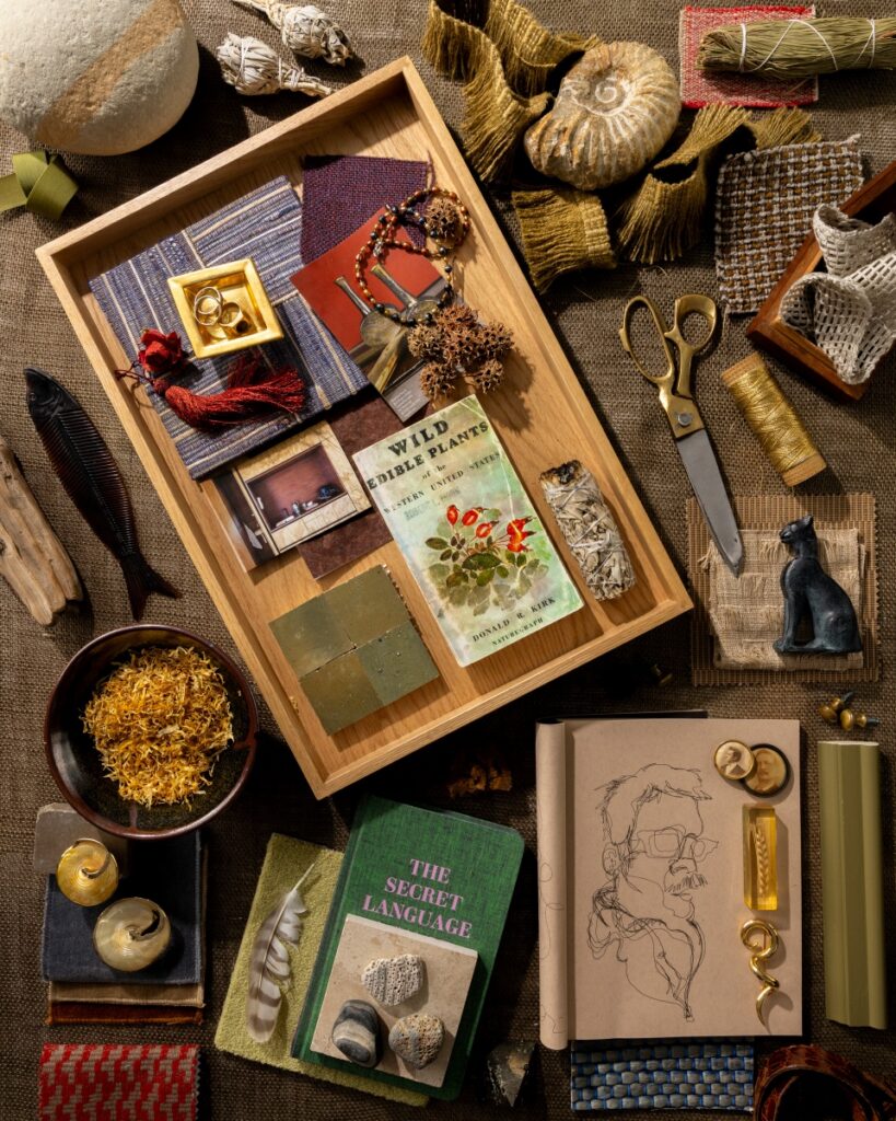 A creative mood board by Danny Mankin featuring a collection of vintage books, personal sketches, gold jewelry, and varied fabric textures laid out on a burlap surface.