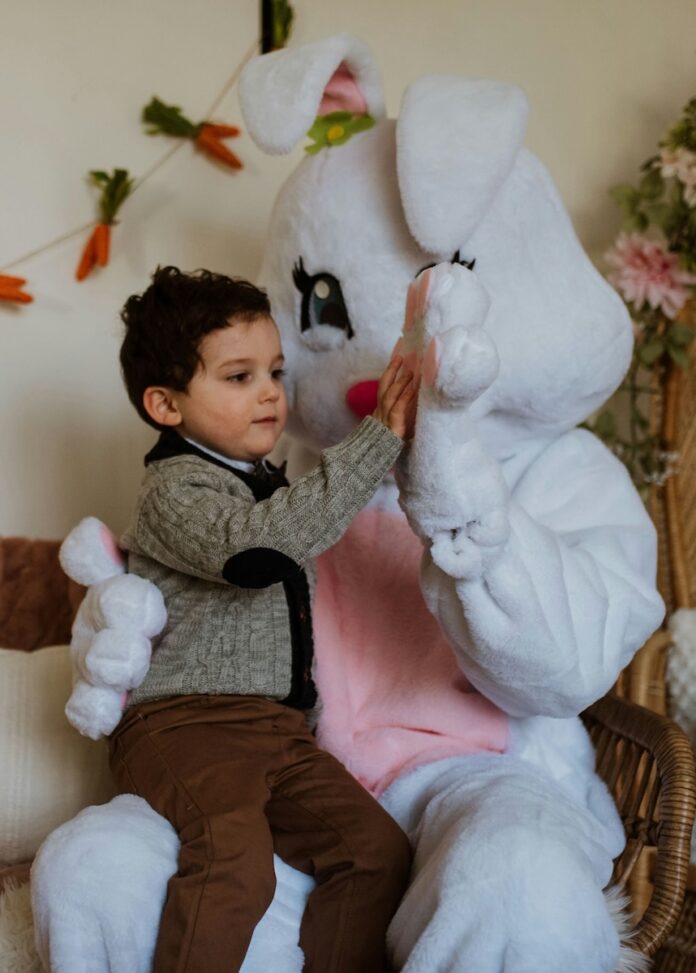 An Easter Bunny high fives a kid sitting on its lap for a picture.