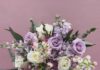 Easter Florists for Your Table Centerpieces and Beyond A florist holds a bouquet of pastel purple and white flowers in front of a purple background.