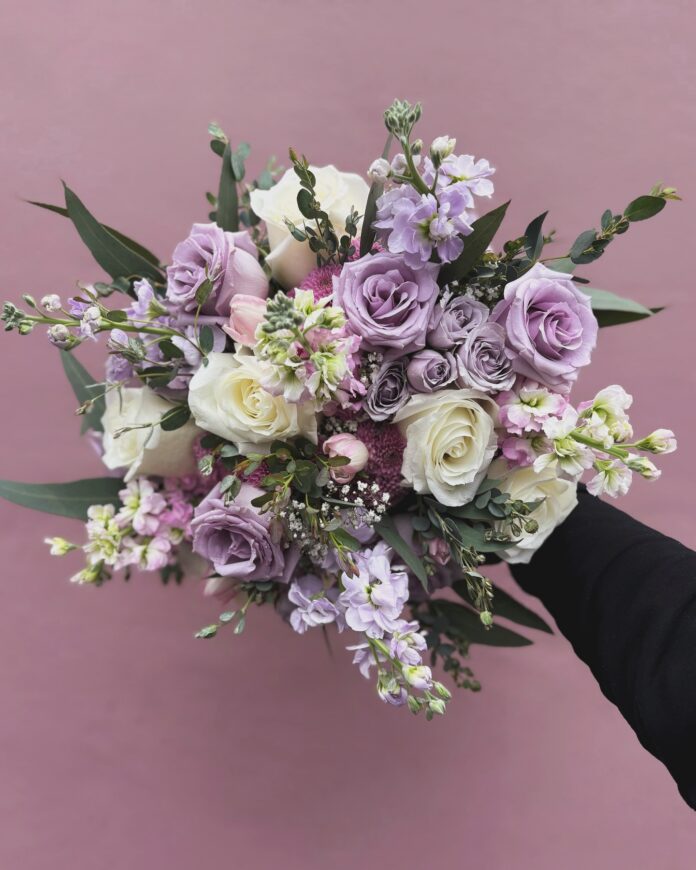 A florist holds a bouquet of pastel purple and white flowers in front of a purple background.