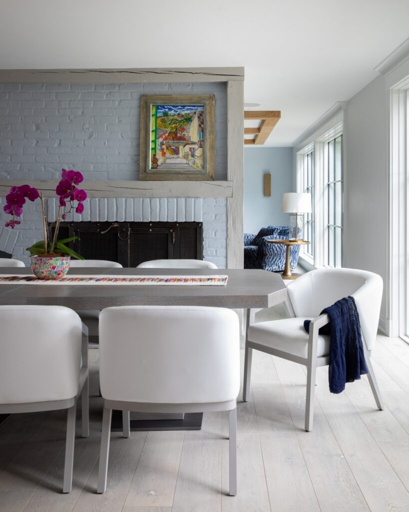 A bright dining area in a Fox Chapel farmhouse featuring white modern chairs, a light wood floor, and a light blue brick fireplace decorated with colorful artwork.