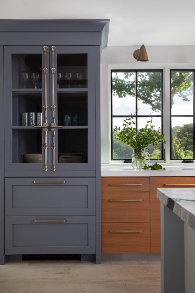 A sleek, modern kitchen renovation in a Fox Chapel farmhouse featuring custom blue cabinetry with brushed brass hardware, natural wood drawers, and a large window overlooking the landscape.