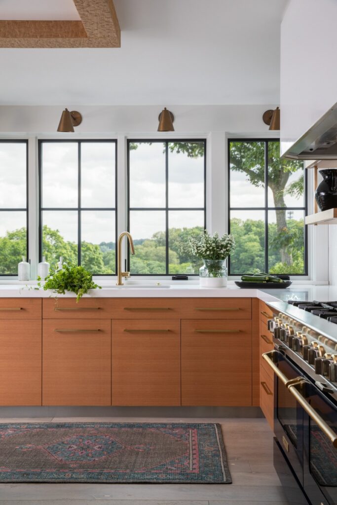 A renovated Fox Chapel farmhouse kitchen featuring natural white oak cabinets with brushed brass hardware, a vintage-style area rug, and Kolbe Vista Luxe windows overlooking a picturesque landscape.