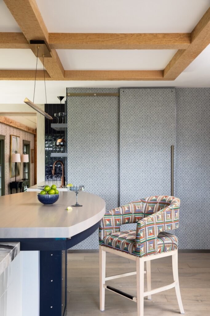 A contemporary breakfast bar in a Fox Chapel farmhouse featuring a light wood countertop, a colorful geometric pattern barstool, and a sliding gray patterned pantry door under exposed oak beams.