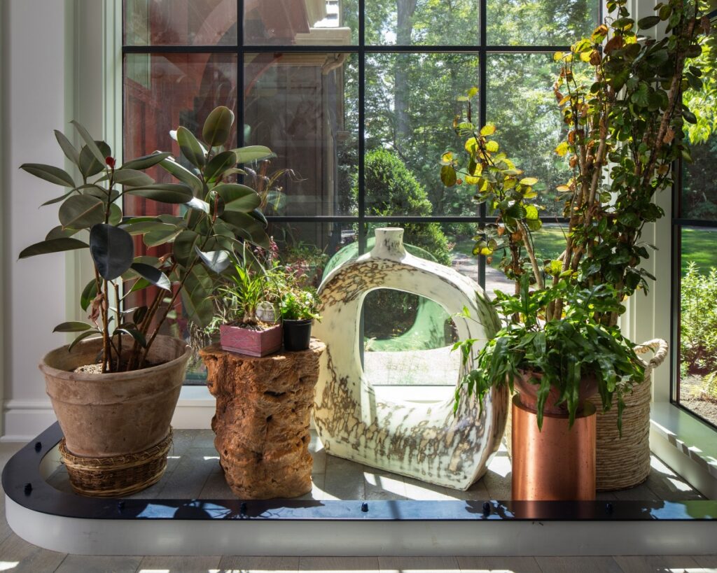 A bright indoor plant corner in a Fox Chapel farmhouse renovation featuring floor-to-ceiling Kolbe Vista Luxe windows, a large sculptural vase, and lush greenery.