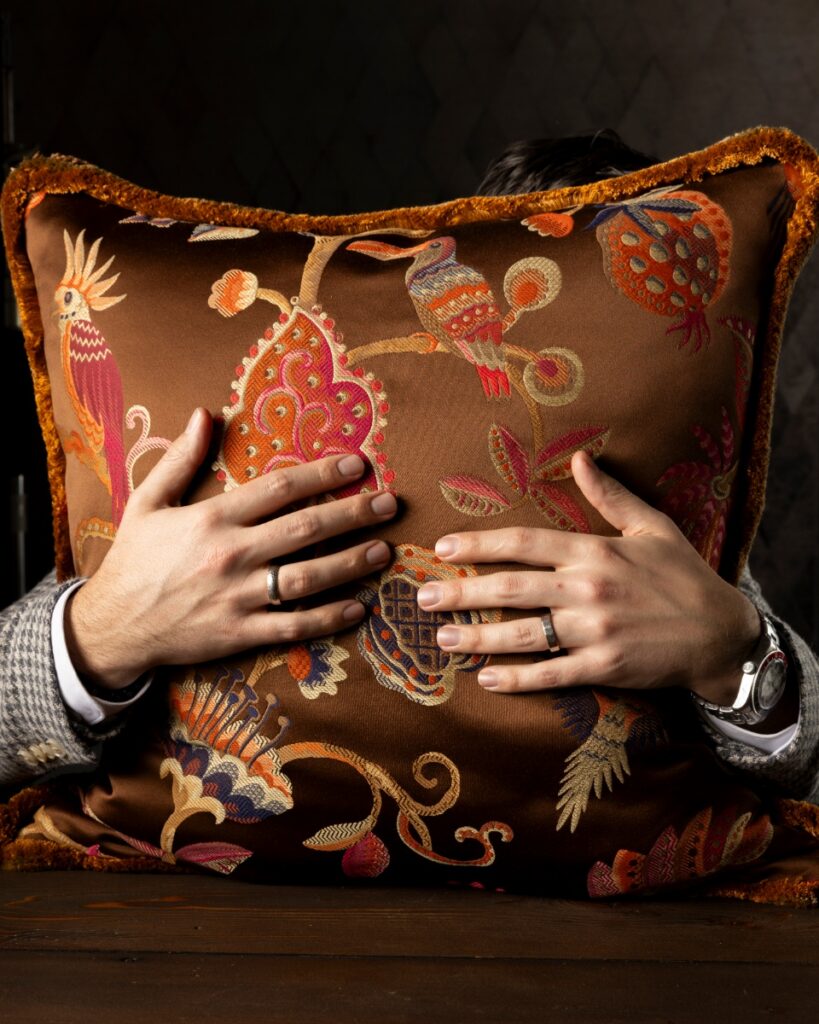 A luxury throw pillow by Brenda Friday Design featuring Schumacher’s Birds of Paradise fabric in Chestnut, with intricate embroidered birds and floral motifs and a fringed trim.
