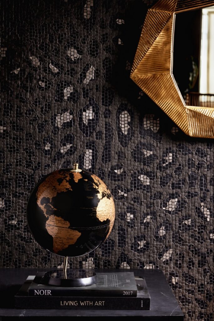 Leo Sea Grey mosaic tile pattern by Splash, featuring a handcrafted leopard spot design in natural stone tesserae, styled with a black and gold globe and a geometric mirror.