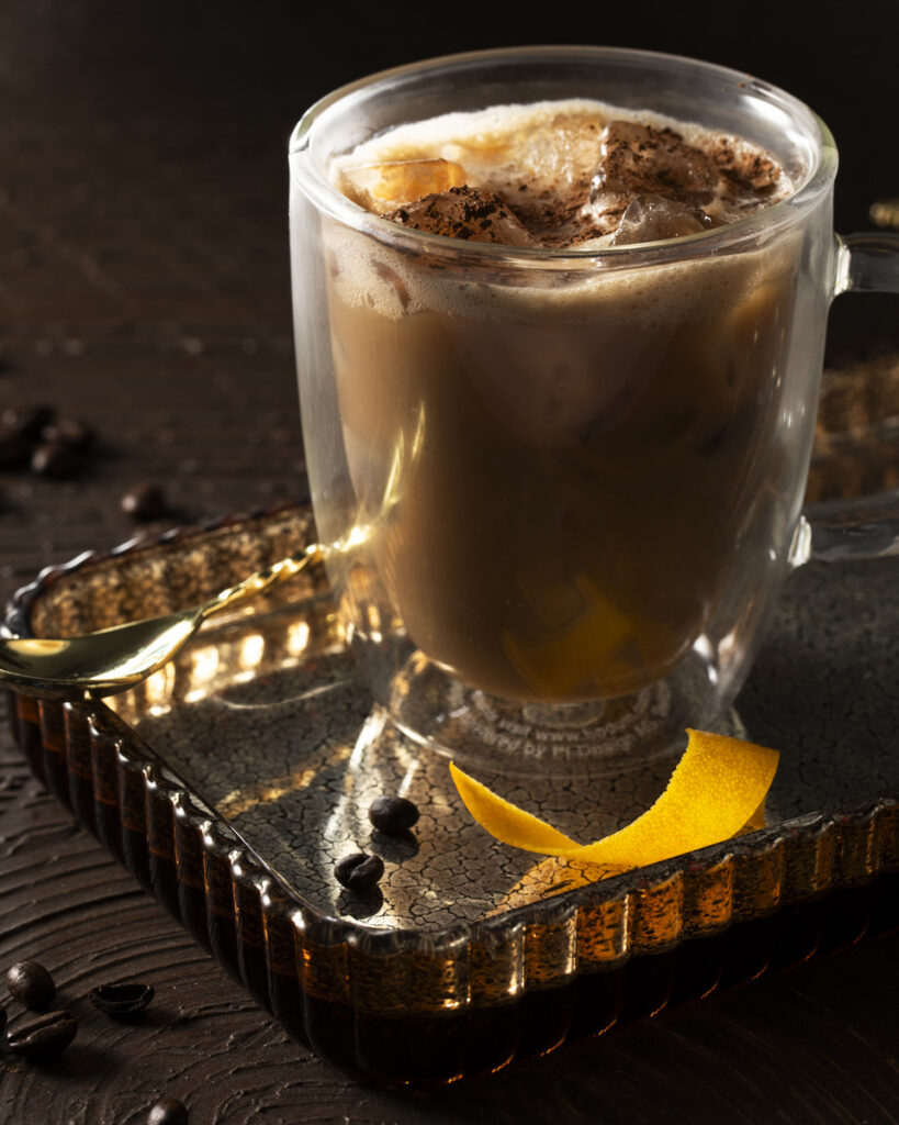 A glass mug filled with a dark, creamy-looking A non-alcoholic Irish coffee garnished with a lemon peel and what appears to be coffee beans or espresso grounds.