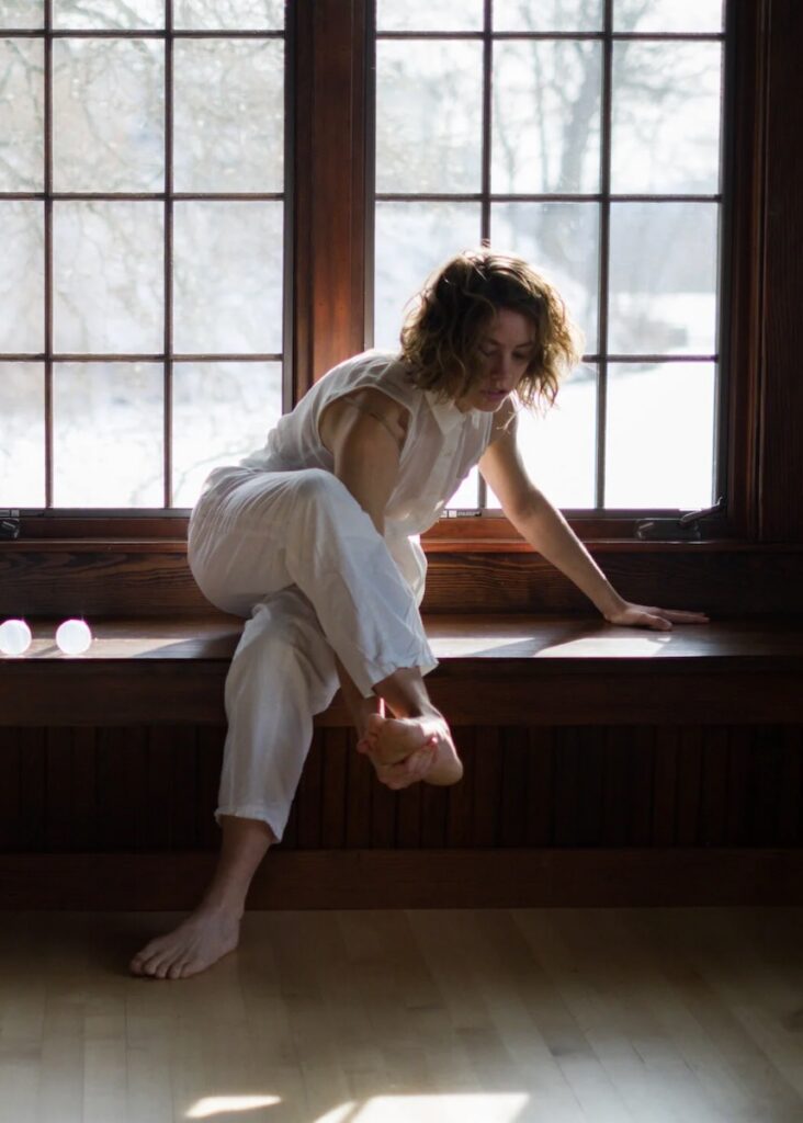 A woman stretches in a white outfit while sitting on a bench in front of sunny windows.