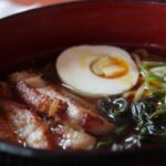 A bowl of hangover ramen with slices of pork, a soft boiled egg, and greens.