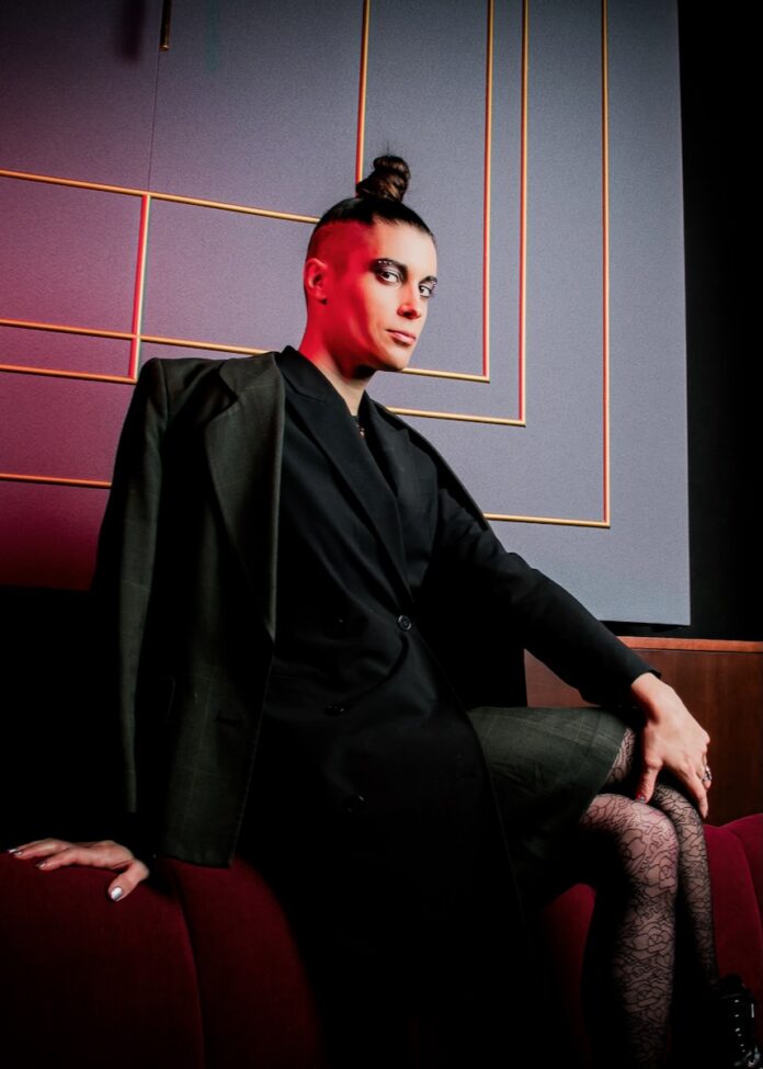 Performer Treasure Treasure as Hedwig in the City Theatre production of "Hedwig and the Angry Inch," wearing a voluminous blonde wig and a colorful, frayed denim patchwork outfit.