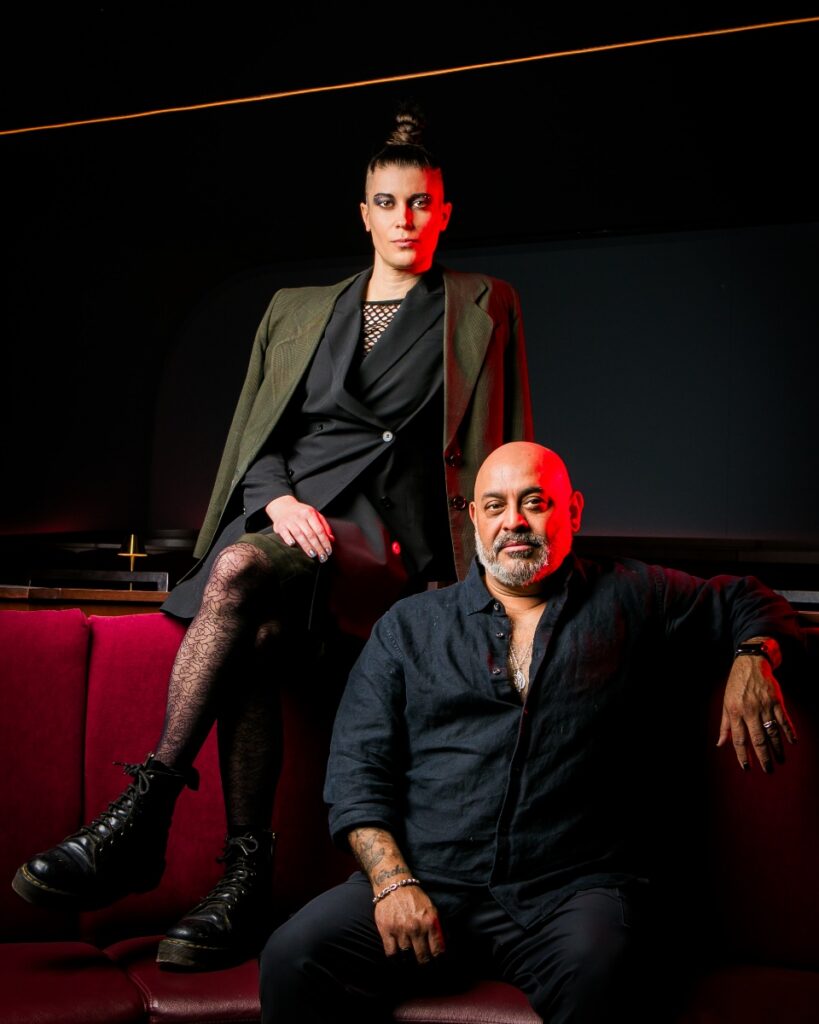 A dramatic close-up of Treasure Treasure as Hedwig, featuring heavy glitter eye makeup, bold red lipstick, and an oversized blonde wig with feathered layers.