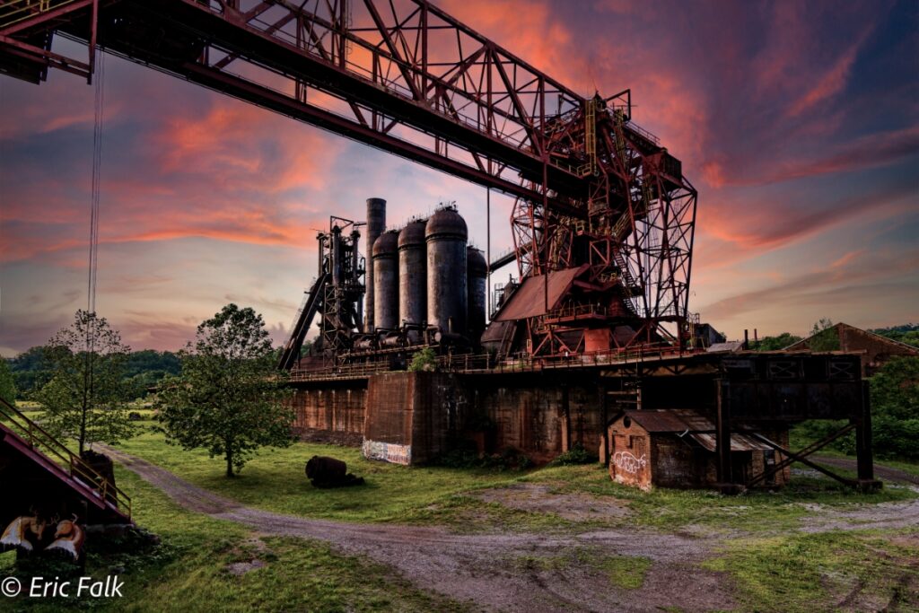 A sunset view of the historic Carrie Blast Furnaces in Homestead, featuring the industrial steel structures and a large overhead crane against a vibrant orange and purple sky.