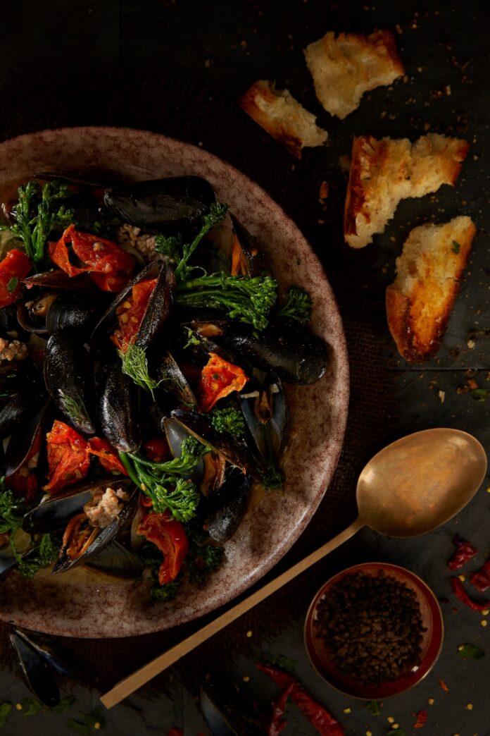 A bowl of steamed black mussels with broccolini, sun-dried tomatoes, and savory ground sausage served with toasted bread at Eighty Acres Kitchen & Bar in Plum.
