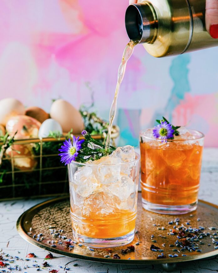 Two short cocktail glasses hold a Hippity rose hip and lavender easter cocktail with small purple flower garnishes and a basket of eggs sitting in the background.