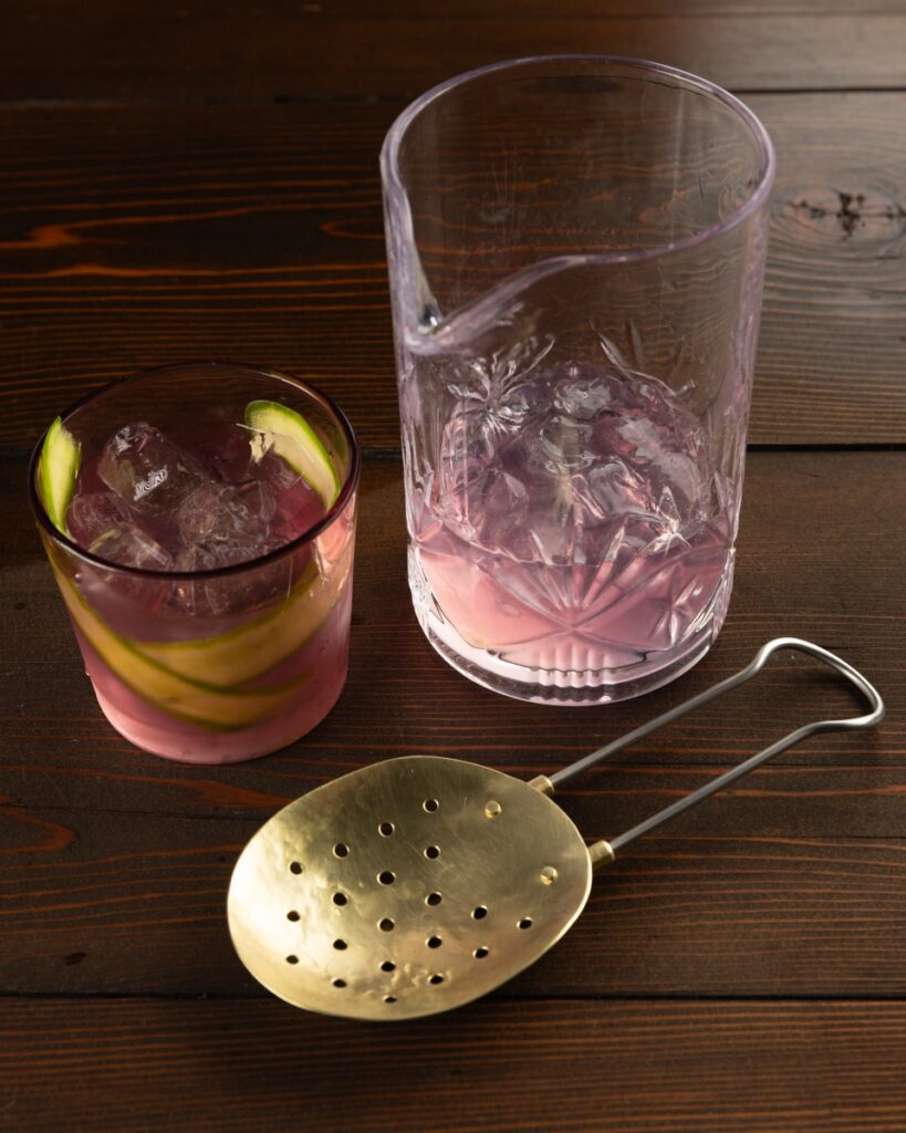 Handcrafted Studebaker Metals brass cocktail strainer with a hammered finish, displayed on a dark wood table next to a crystal mixing glass and a pink cocktail with cucumber garnishes.