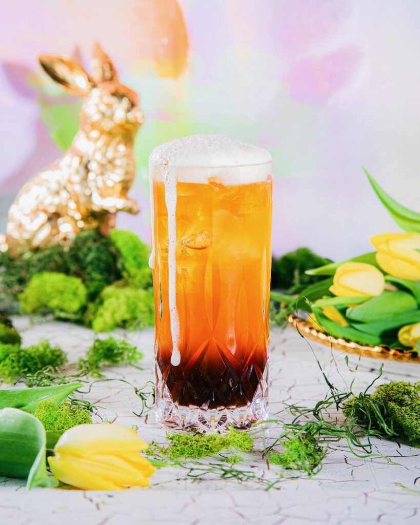 A Hoppity IPA Cocktail in a tall beer glass sits on a white table surrounded by Easter grass, eggs, and a gold bunny by the pastel backdrop.