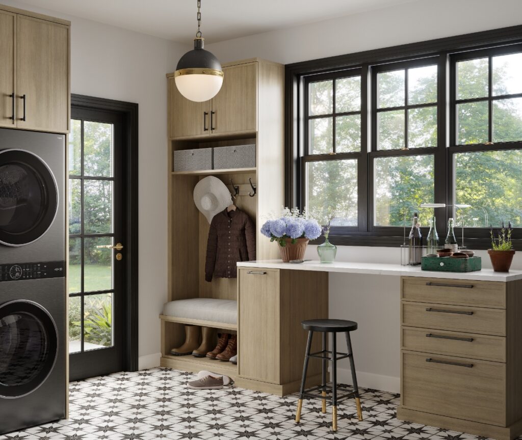 A well-organized laundry room and mudroom featuring light wood cabinetry, a stacked dark gray washer and dryer, a built-in bench, a desk area, and a patterned tile floor.