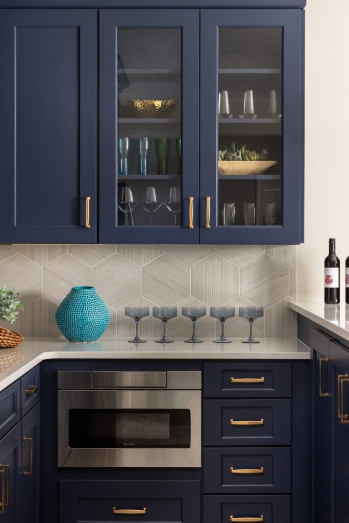 A modern home bar and kitchenette featuring navy blue cabinetry with gold hardware, a neutral hexagon tile backsplash, and a built-in stainless steel microwave.