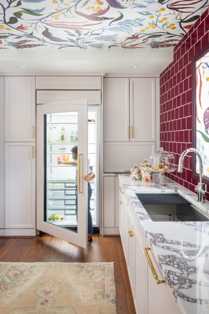 A vibrant butler's pantry designed by Colleen Simonds featuring a floral wallpapered ceiling, bold burgundy subway tile, marble countertops, and a child reaching into a glass-front refrigerator.
