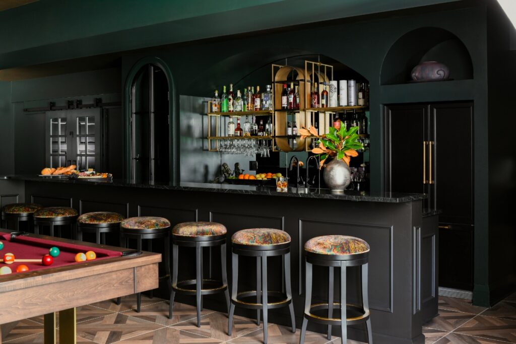 A sleek, full-scale basement bar featuring black cabinetry, a dark marble countertop, patterned swivel barstools, and a pool table in a renovated entertainment space.