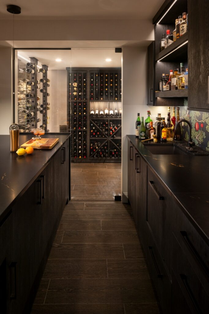 A sleek, modern basement wine cellar and bar area featuring black cabinetry, dark quartz countertops, and a glass-enclosed walk-in wine room with custom wood shelving.