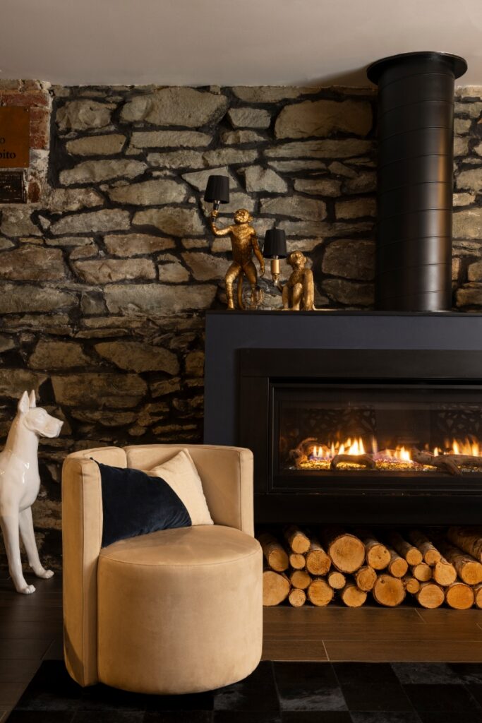 A modern basement seating area featuring a modern black fireplace, a rustic stone accent wall, and a tan swivel chair with velvet pillows.
