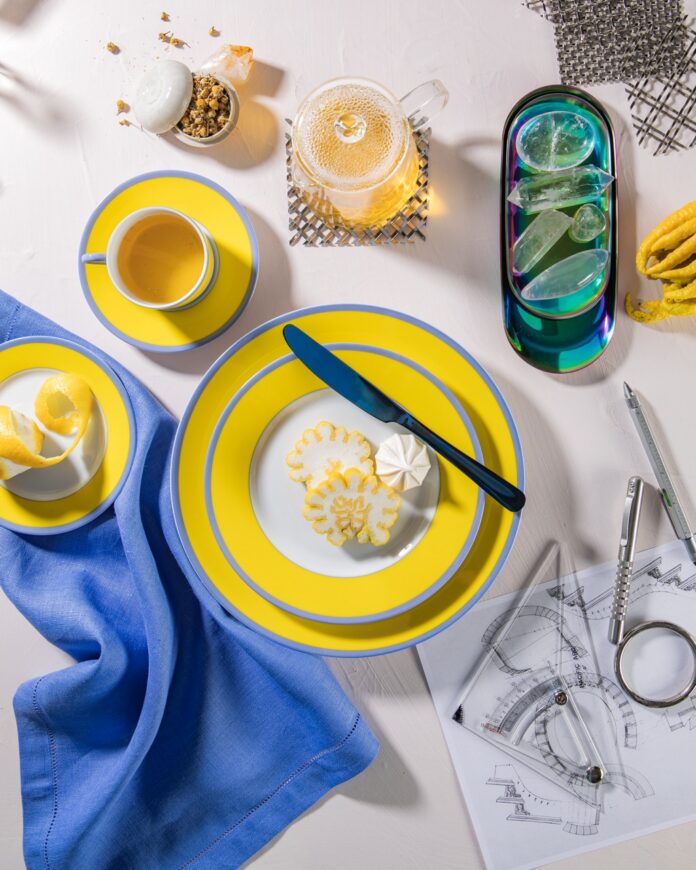 A two stack of yellow plates with blue edges sits on a white table with a blue cloth to the left of it, a yellow saucer plate above it, and then a plate of crystals to the right of it.