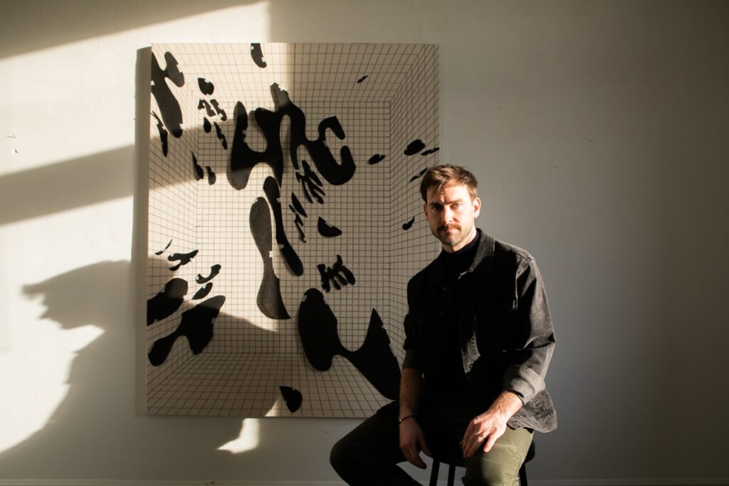 John Burt Sanders sits in front of an abstract collage of black spots.