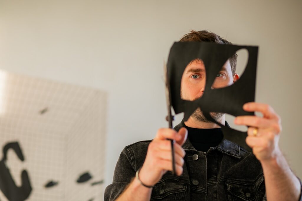 John Burt Sanders cuts out shapes in front fo his face from a black sheet of paper.