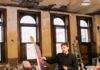 John Burt Sanders Explores Color and Abstraction in Art Artist John Burt Sanders standing in his Wilkinsburg studio next to a large abstract painting featuring textured fields of pink, blue, and yellow inspired by Rococo aesthetics and video game graphics.