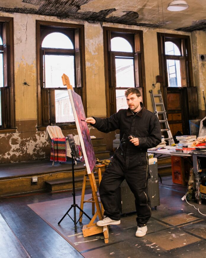 Artist John Burt Sanders standing in his Wilkinsburg studio next to a large abstract painting featuring textured fields of pink, blue, and yellow inspired by Rococo aesthetics and video game graphics.