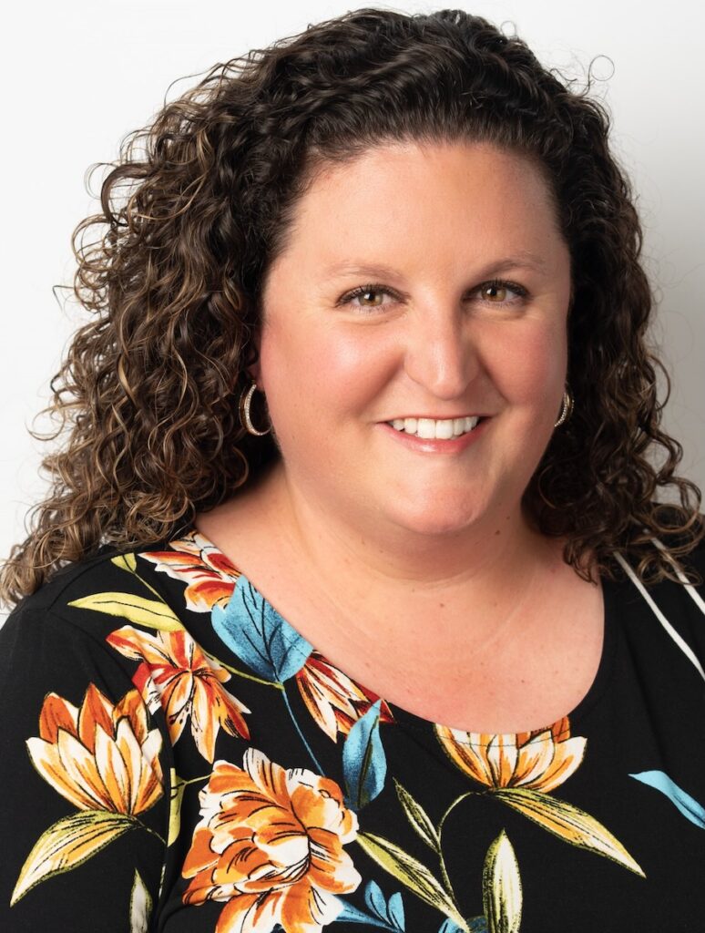 A woman with brown curls and a floral black shirt on.