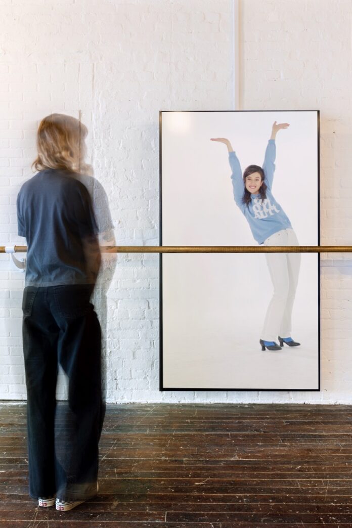 A person stands in front of a barre bar as another person poses on a screen behind it.